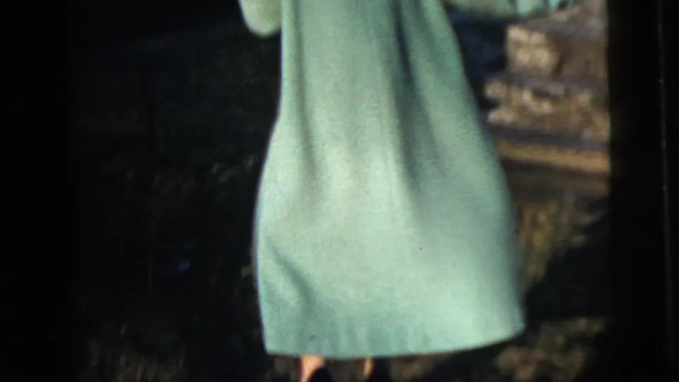 Back view of a woman in a pale green coat near stone steps, with heavy shadow and warm 8mm color from the 1958 New York City clip.
