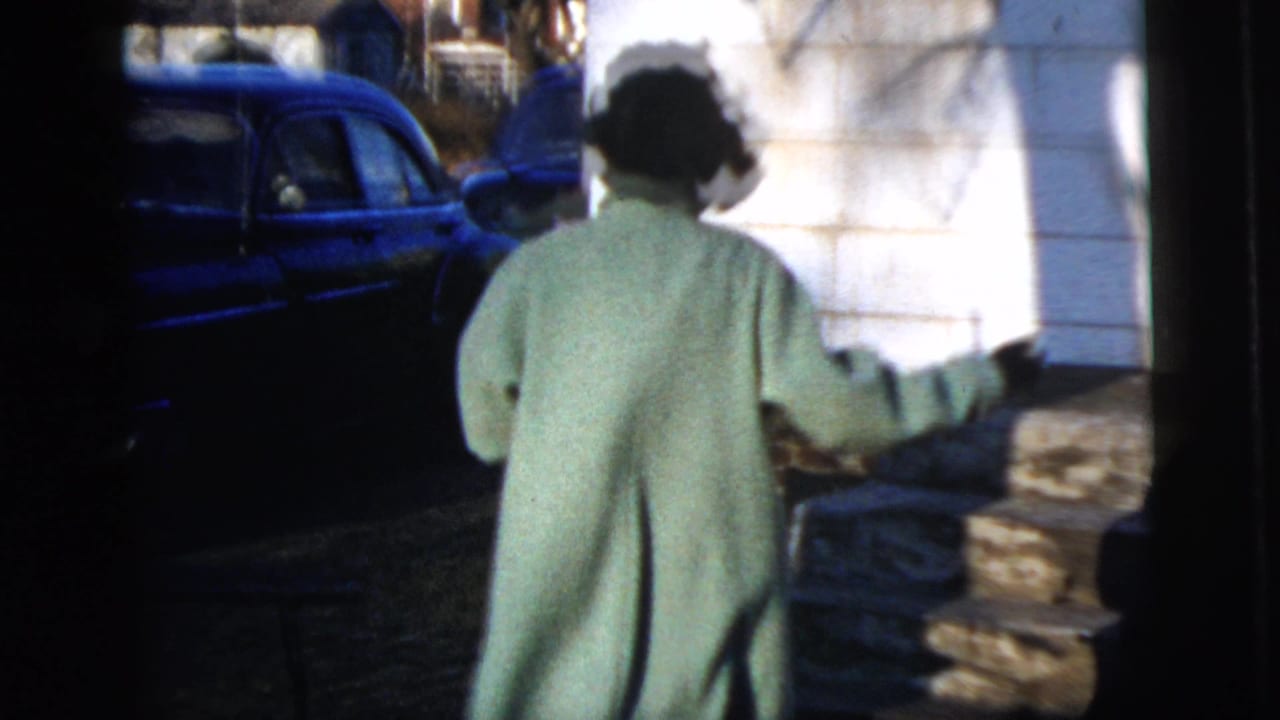 In 1958 New York City home-movie footage, a woman in a long mint-green coat walks toward a parked blue sedan on a residential street.