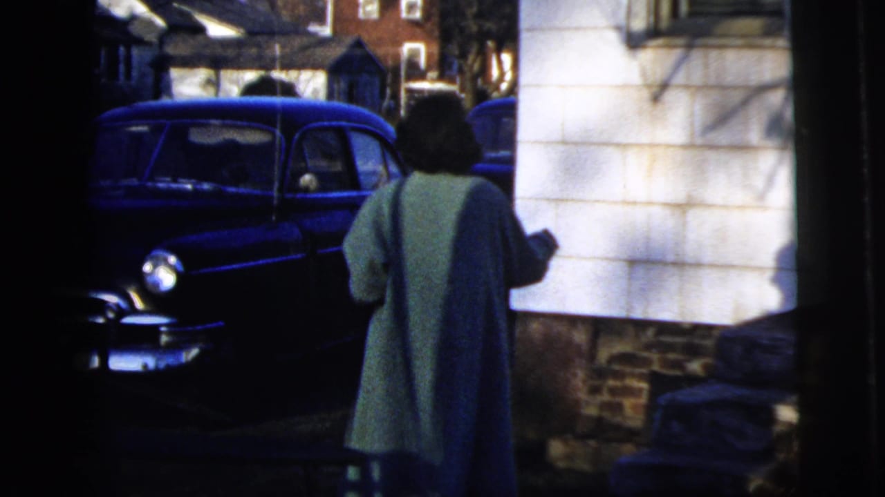 The woman continues toward the parked blue sedan, seen from behind near white siding and a residential foundation in New York City footage.
