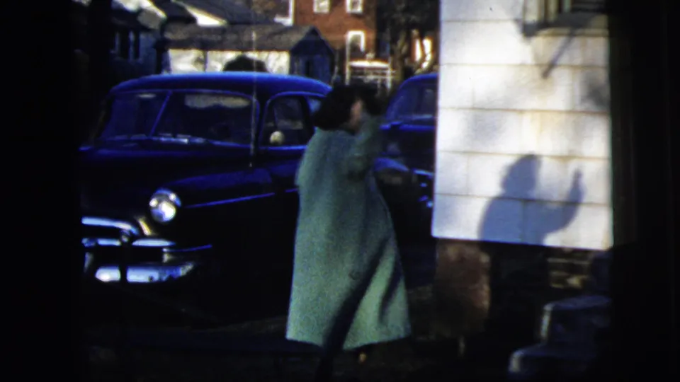 The woman turns back and appears to wave toward the camera beside the blue sedan on a residential New York City street.