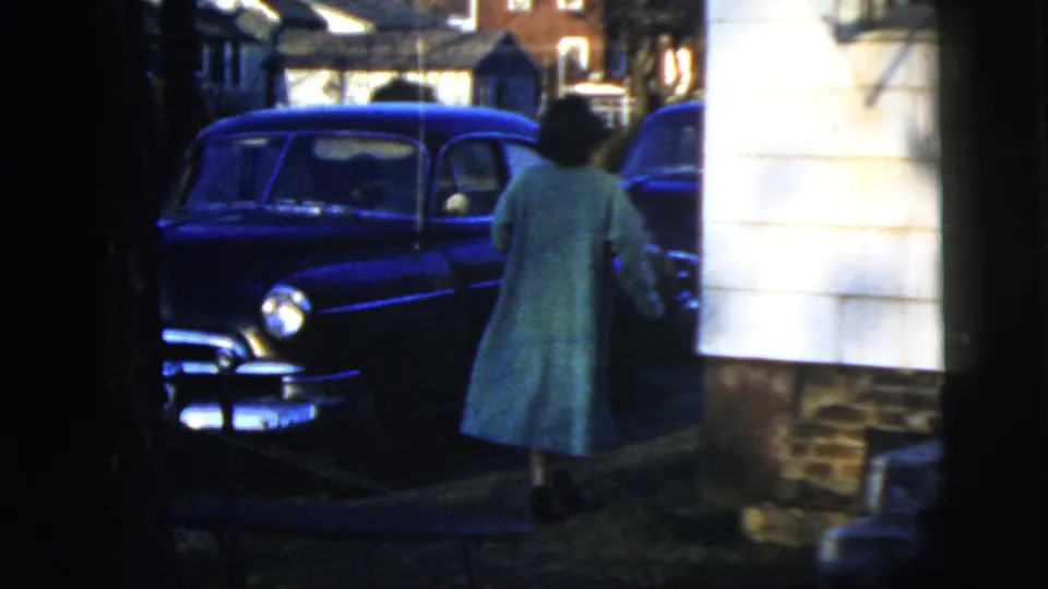 The woman moves along the side of the house near the parked blue sedan, with strong shadows across the 1958 home-movie frame.