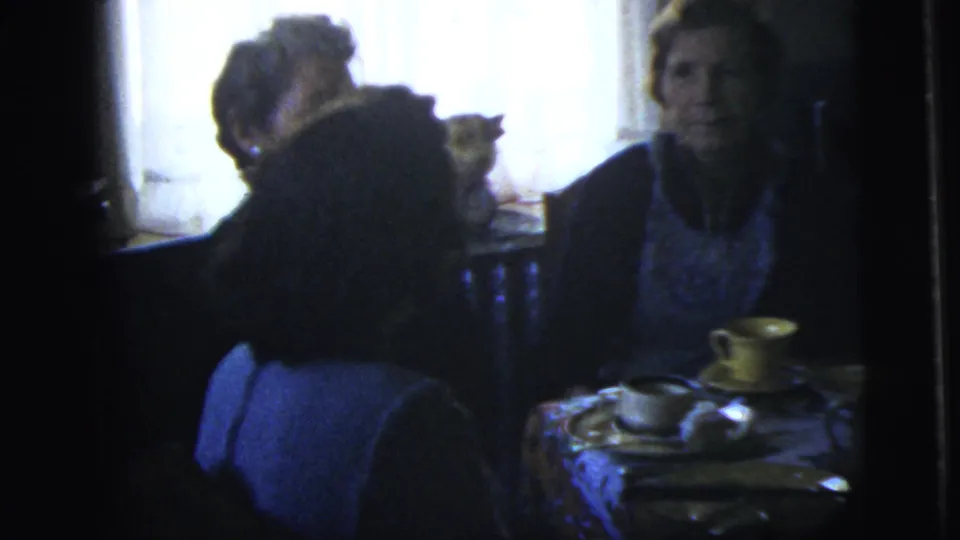 1958 New York City home-movie frame showing adults seated at a small dining table with cups, food, and a bright curtained window.