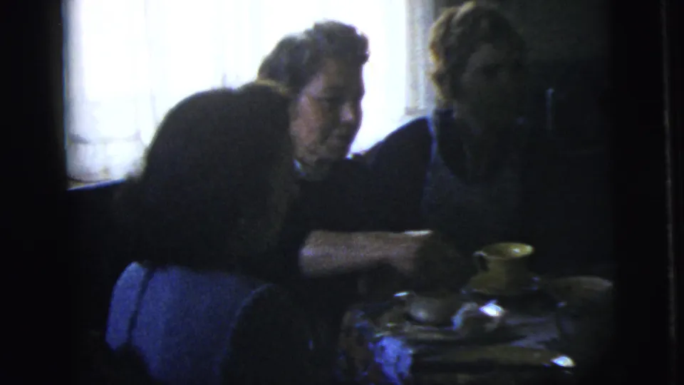1958 New York City home-movie frame with adults at a table while one person reaches across dishes and yellow cups.