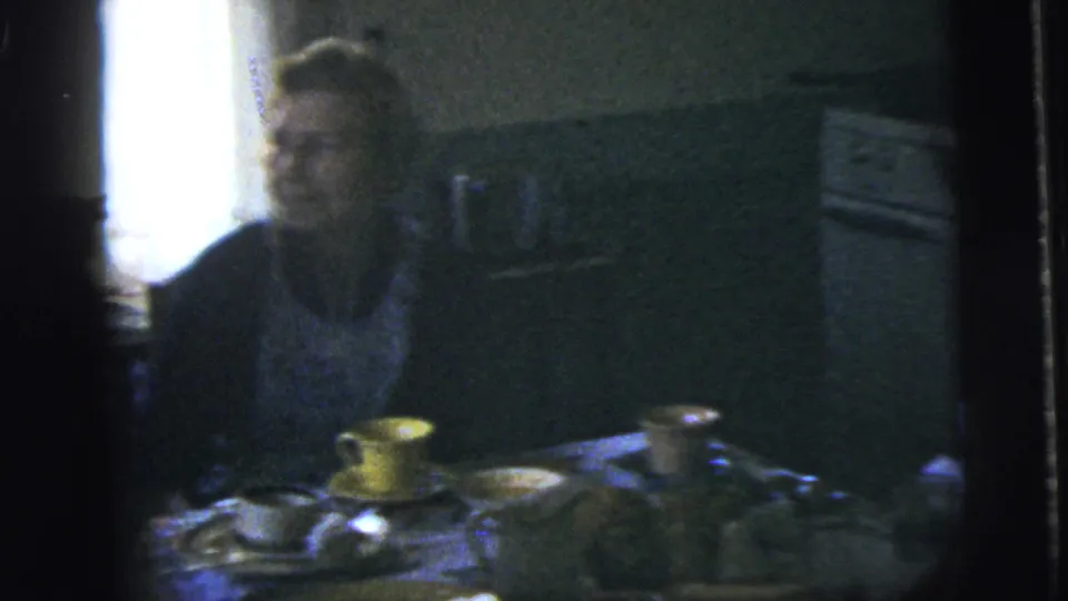 1958 New York City home-movie frame showing a seated woman beside a table with cups, dishes, and food in a private interior.