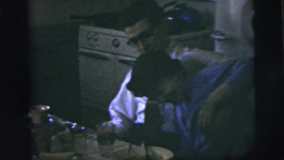 1958 New York City home-movie frame showing adults leaning near a table with cups, dishes, and kitchen cabinetry in view.