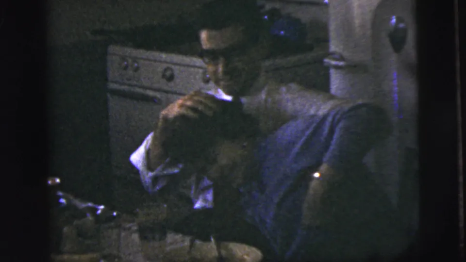 In a dim 1958 New York City kitchen, a man leans over a woman near a stove, dishes, and small table items.