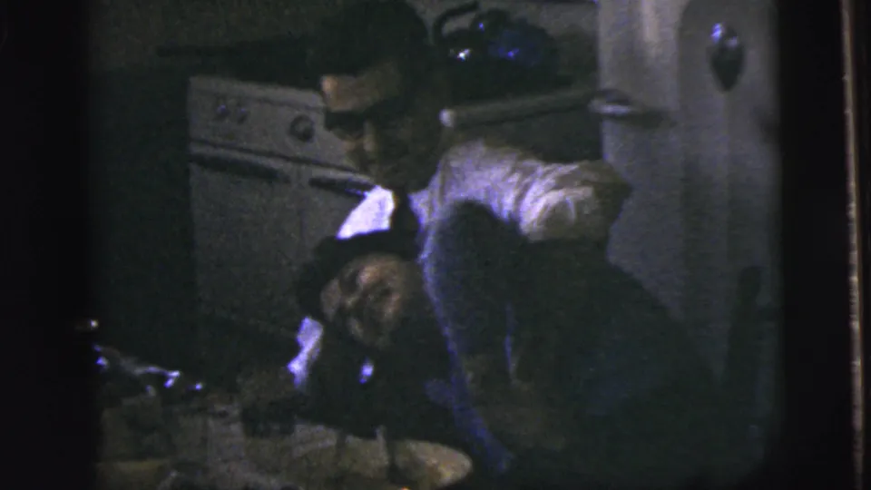 The woman leans back and laughs or reacts as the man bends close beside a stove in the dim kitchen setting.