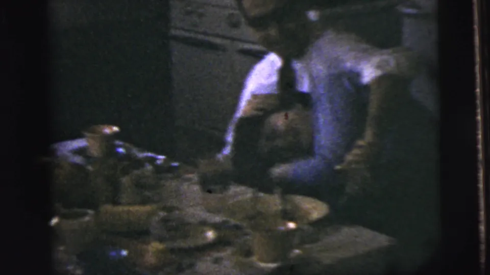 A man in a light shirt bends over a woman near dishes, a bottle, and an older kitchen appliance.
