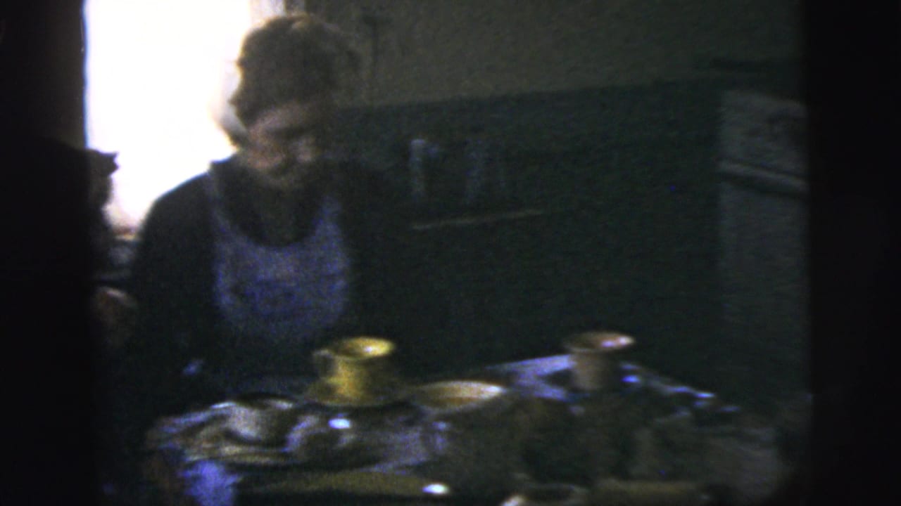 A woman sits at a dim kitchen table in 1958 New York City home-movie footage, with cups and tableware visible.