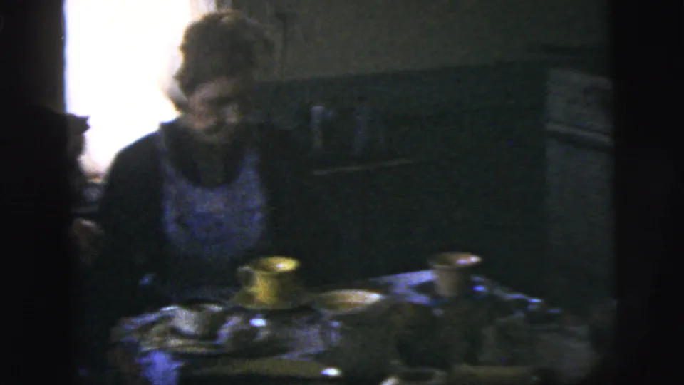 A woman sits at a dim kitchen table in 1958 New York City home-movie footage, with cups and tableware visible.