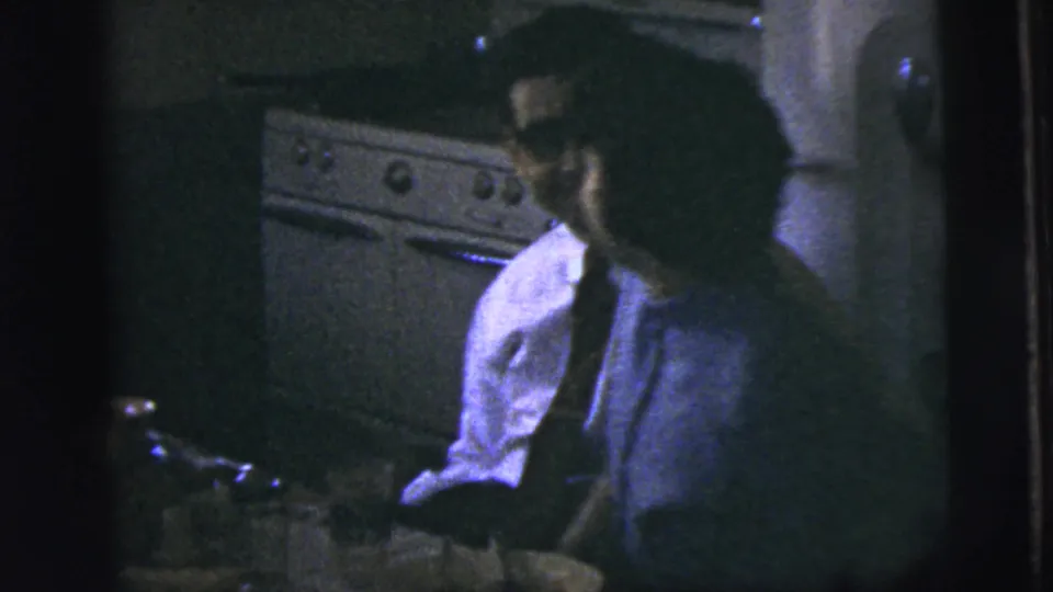 A man in a shirt and tie sits near a kitchen table in 1958 New York City home-movie footage.