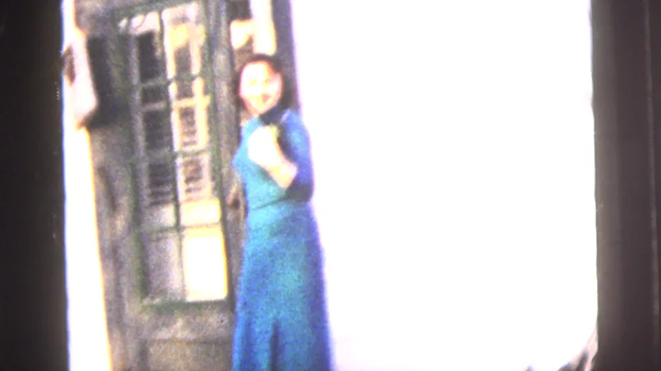 1958 New York City home-movie frame showing a woman in a blue dress standing near a doorway, with the background overexposed.