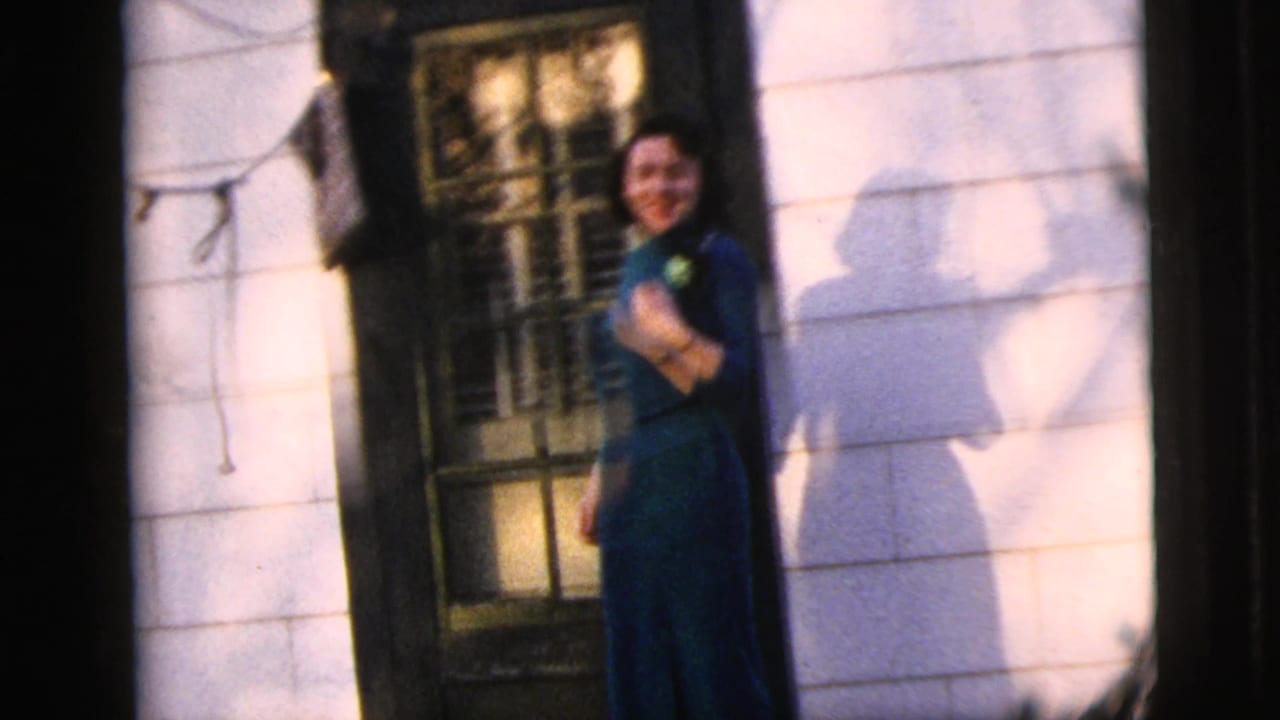 Young woman in a teal dress stands near a house doorway beside white siding and a shuttered window.