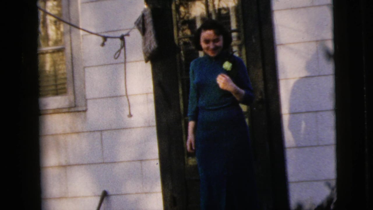 Young woman in a teal dress smiles in a doorway with white siding and a window behind her.