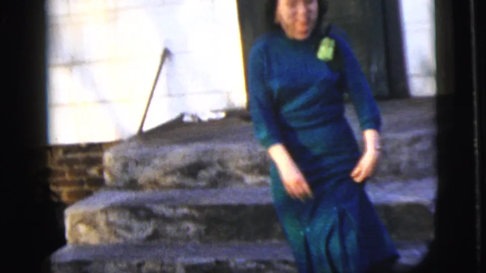 1958 New York City home-movie frame of a young woman in a teal dress walking down stone steps from a house entrance.