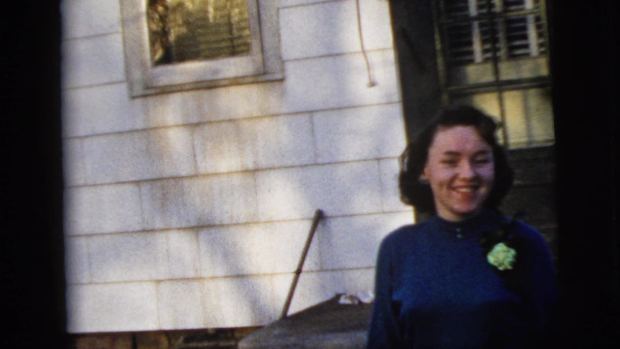 Smiling woman in a blue dress with a green corsage outside a white house.