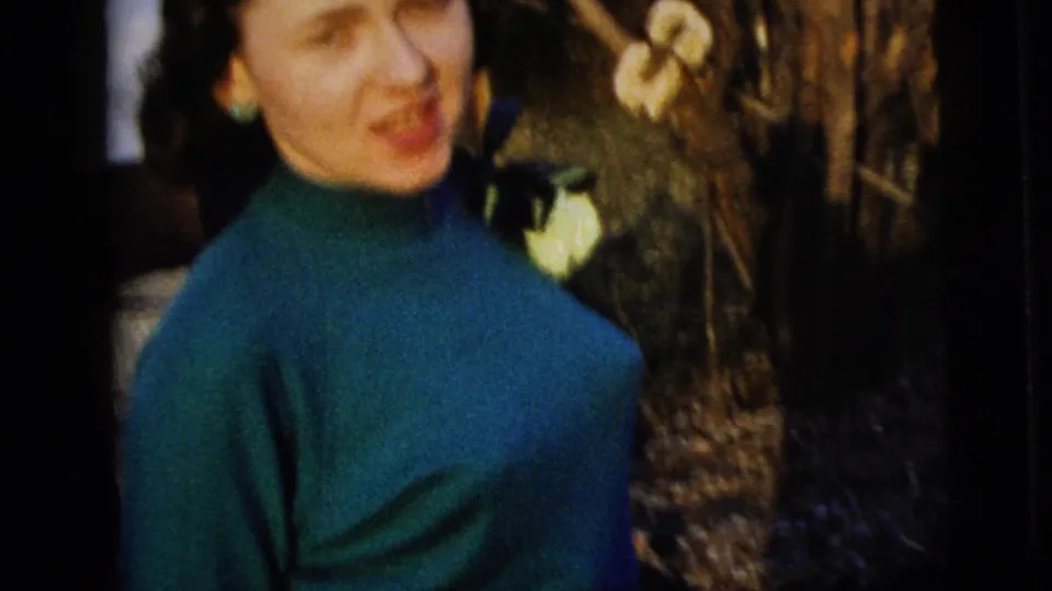 Close handheld 8mm view of the woman in a blue dress and corsage, with garden branches beside the house.