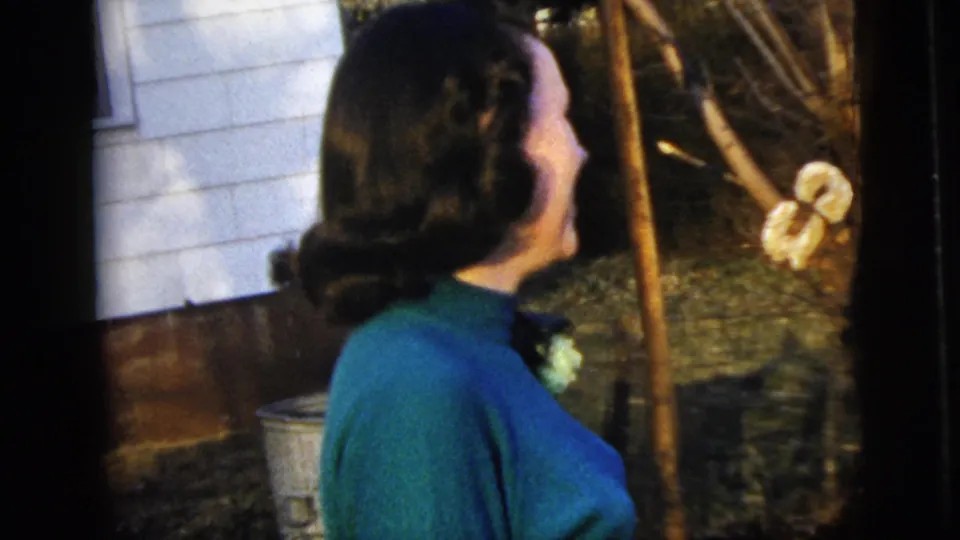 Side view of the woman in a blue dress and corsage facing garden branches near the white-sided house exterior.