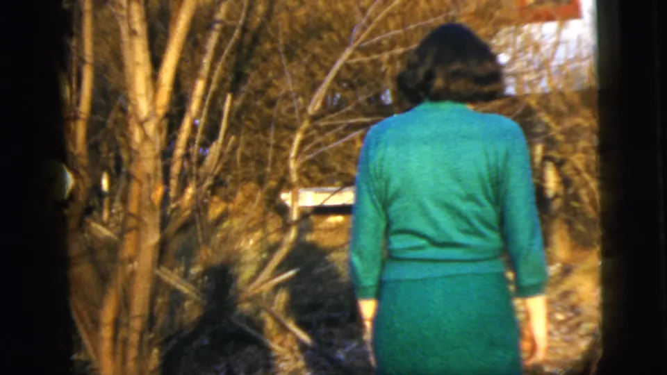 A woman in a green dress walks away from the camera through a leaf-strewn wooded area in New York City home-movie footage.