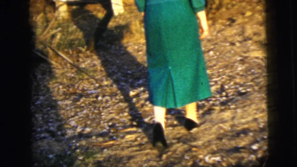 The woman is framed from behind and below as she walks in dark heels along a leaf-covered path in a wooded outdoor setting.