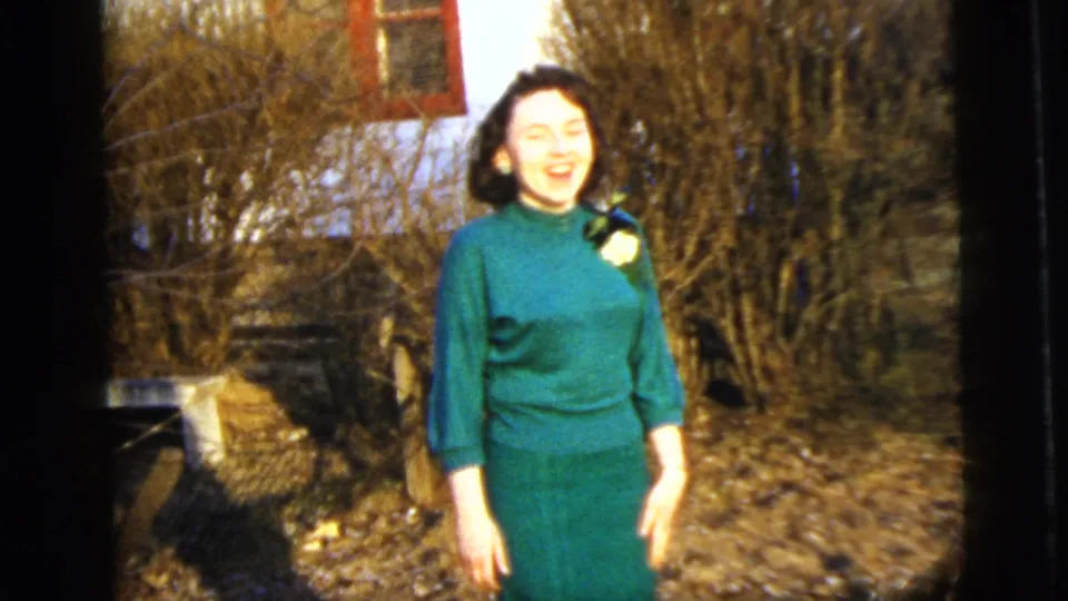 Smiling adult woman in a green dress with a white flower pinned near her shoulder stands in a 1958 New York City garden.
