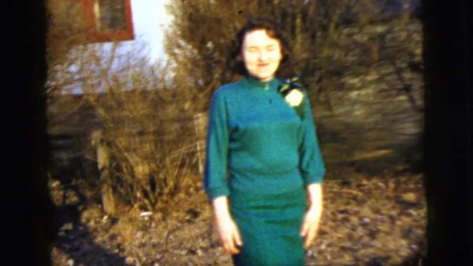 Adult woman in green poses facing the camera in a leaf-strewn New York City garden, with soft 8mm color and frame edges.