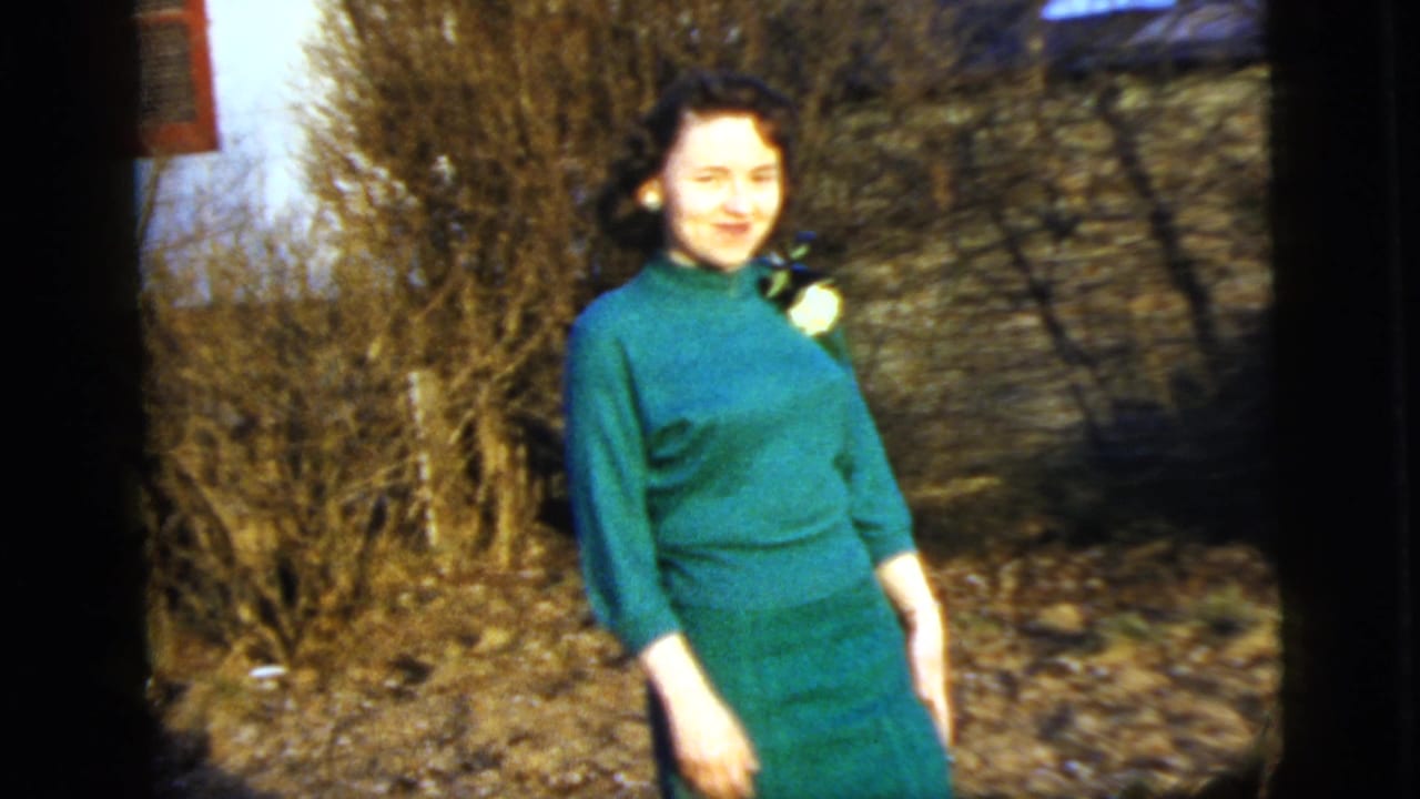 Young woman in a green dress smiles outdoors beside bare shrubs.
