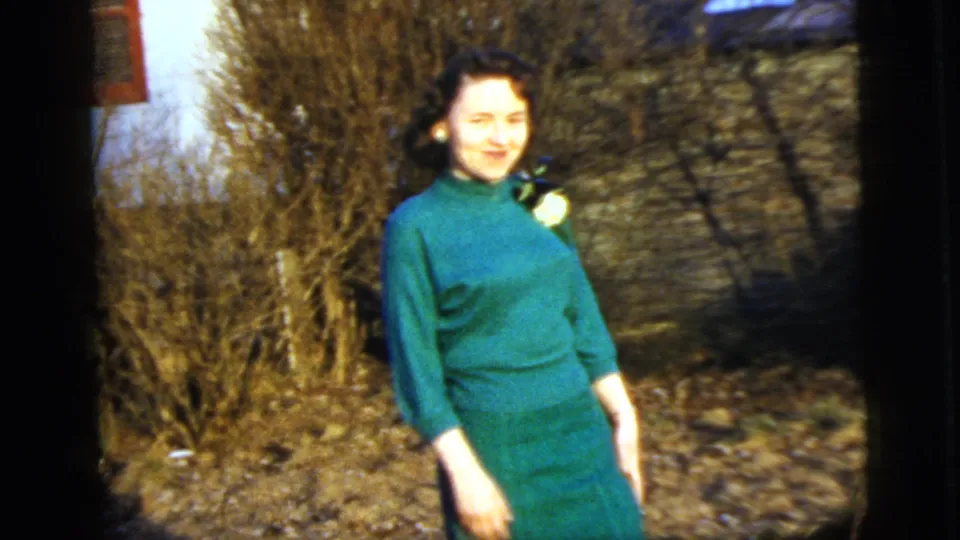 In a 1958 New York home movie frame, a young woman in a green dress with a white corsage smiles outdoors near bare shrubs.
