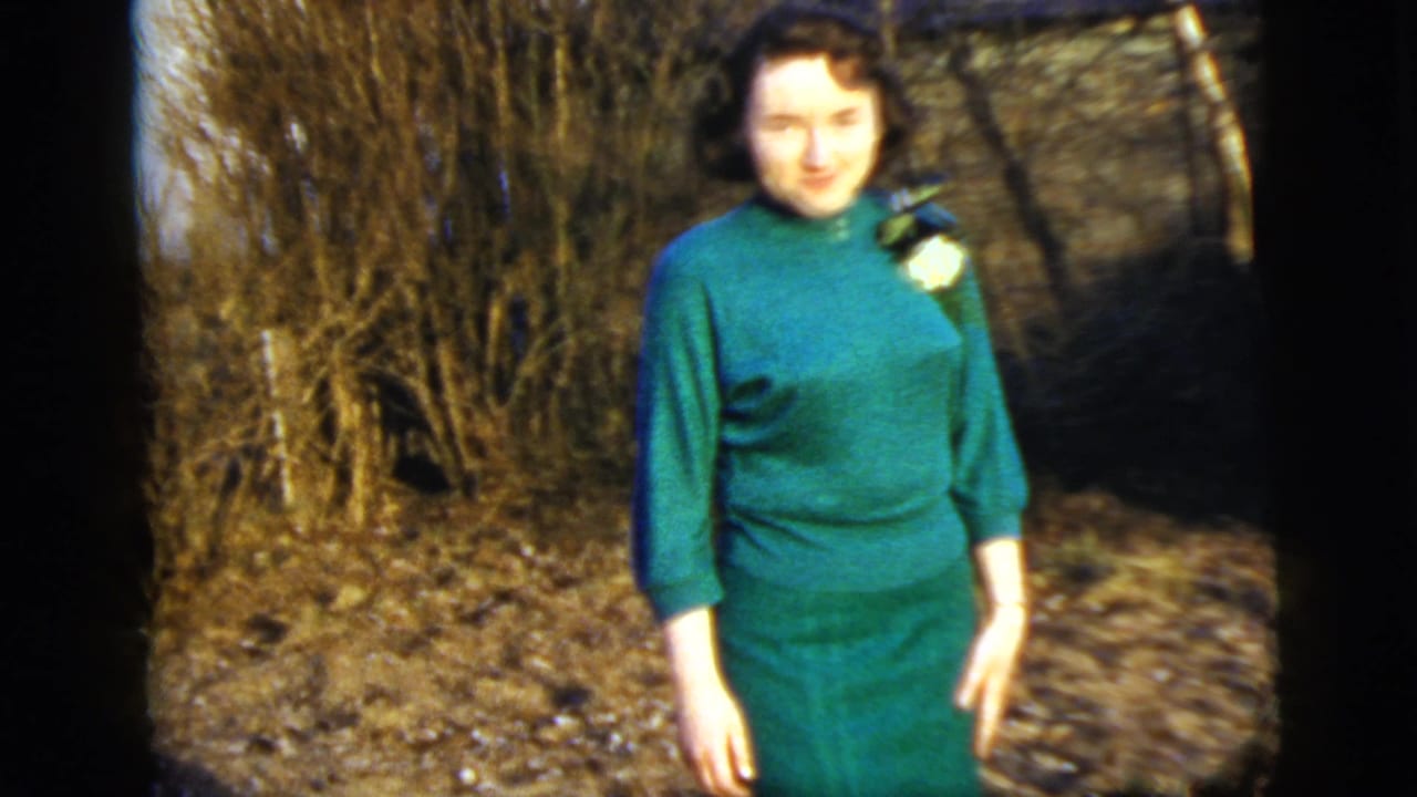 The woman remains posed outside in her green dress and corsage, with bare shrubs and a partial house exterior behind her.