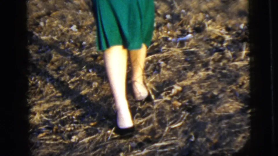 The frame shows the lower half of the posed woman, including her green skirt, legs, and light shoes on uneven outdoor ground.