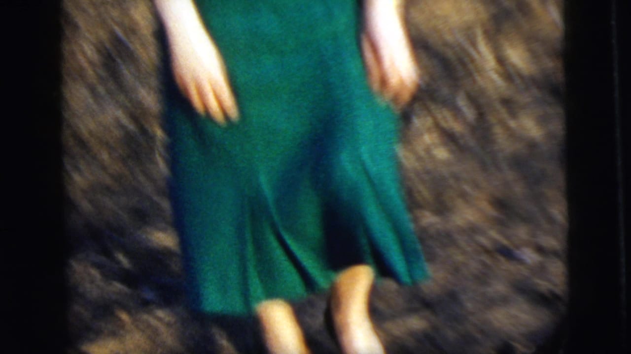 1958 8mm frame of a woman in a green dress standing in dry grass, cropped so her face is out of view.