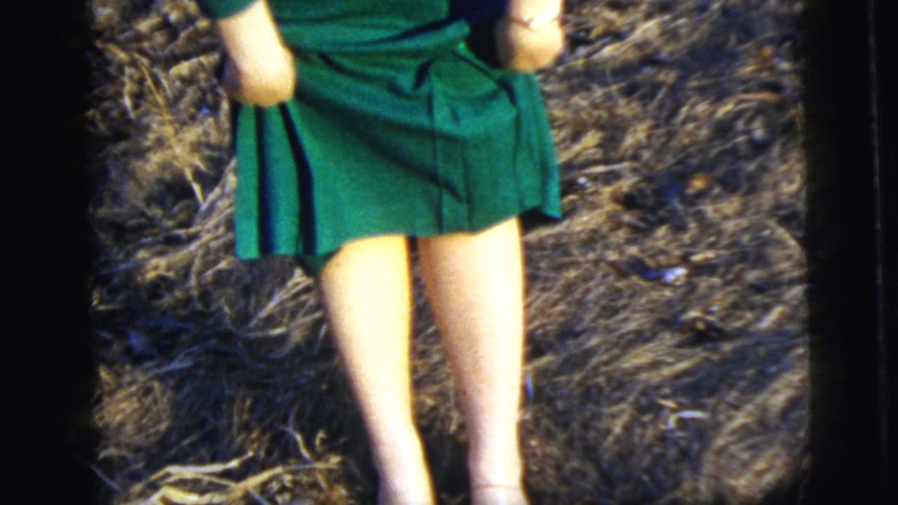 Woman in a green dress lifting the hem slightly while standing on dry grass.