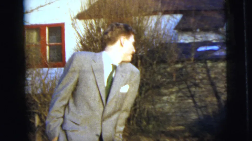 1958 home-movie frame of a man in a gray suit and dark tie outdoors near a light building with red window trim.