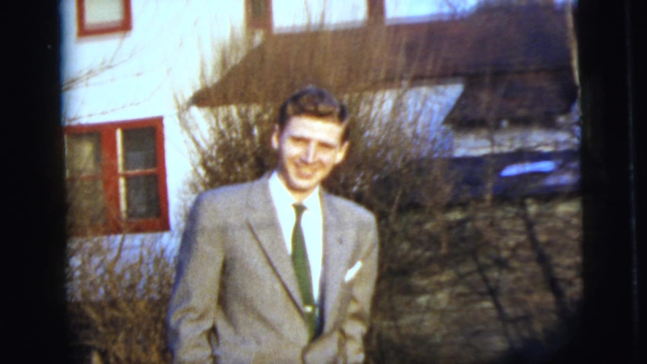 In 1958 New York City home-movie footage, a young man in a light suit and dark tie smiles near a house exterior.