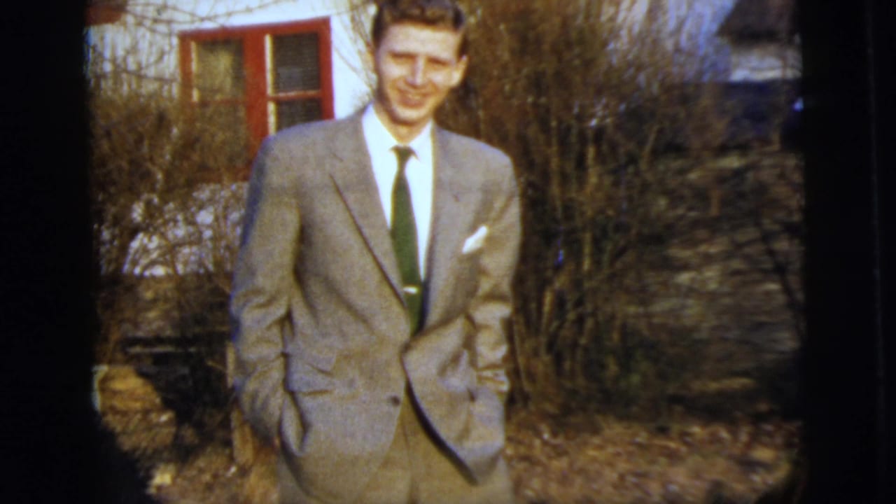 The young man poses in a light suit with hands in his pockets outside a red-trimmed house in archival 8mm footage.