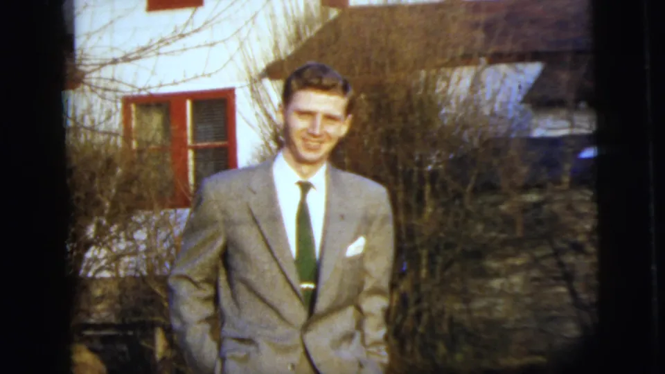 A smiling young man in a light suit remains framed outside the house, with red window trim and winter-bare shrubs behind him.