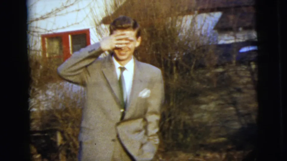 The suited young man playfully covers his eyes with his hand while standing outside the red-trimmed house.