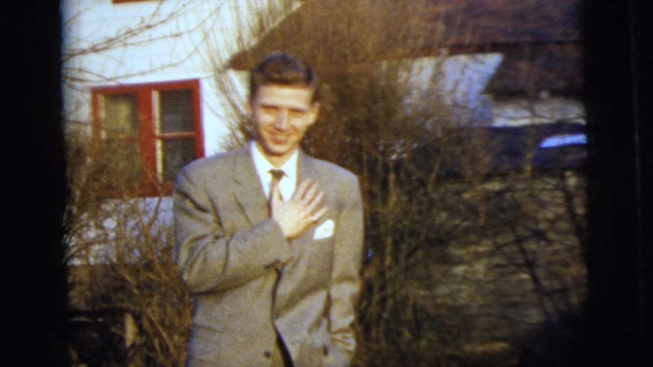 A young man in a light gray suit and tie smiles outdoors in 1958 New York City, with a white building and red-trimmed window behind him.