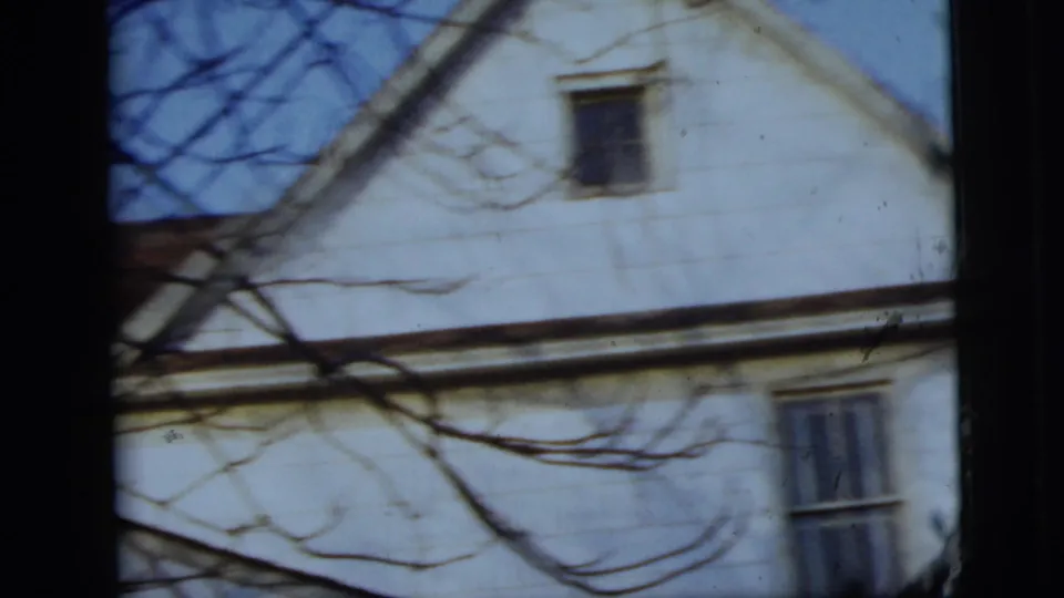 A light-colored house exterior appears behind bare branches, likely part of the same 1958 New York City home-movie scene.