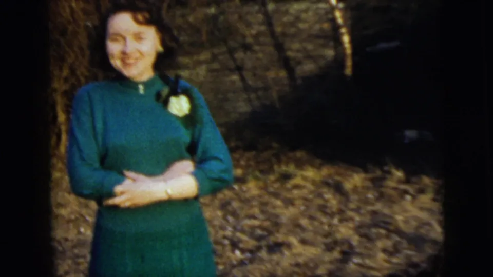 A smiling woman in a green dress with a corsage faces the camera outdoors in the 1958 New York City home movie.