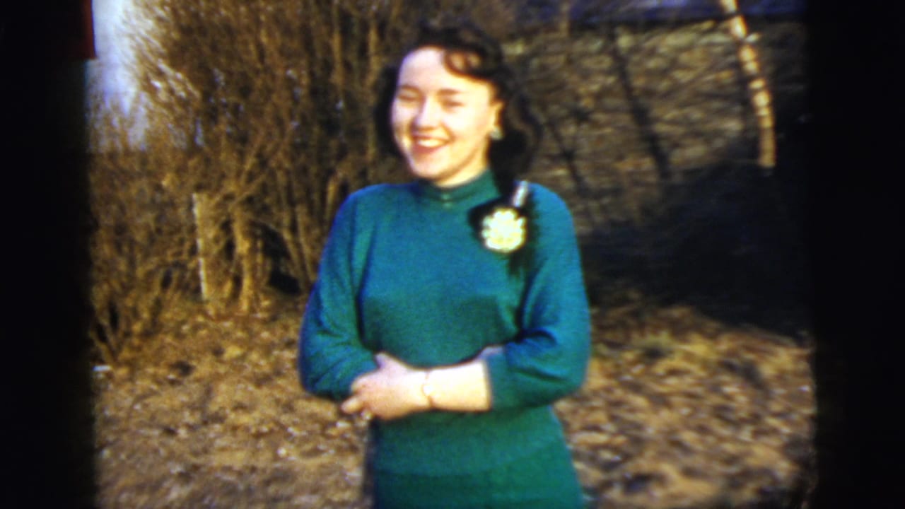 Young woman in a green dress smiles outdoors near leafless trees.