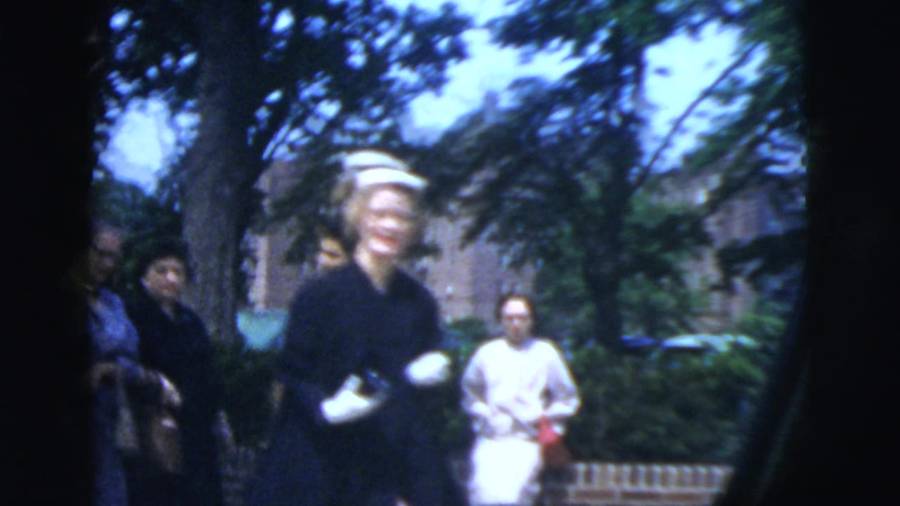 In 1958 New York City home-movie footage, a woman in a white hat and dark dress walks along a shaded residential sidewalk.