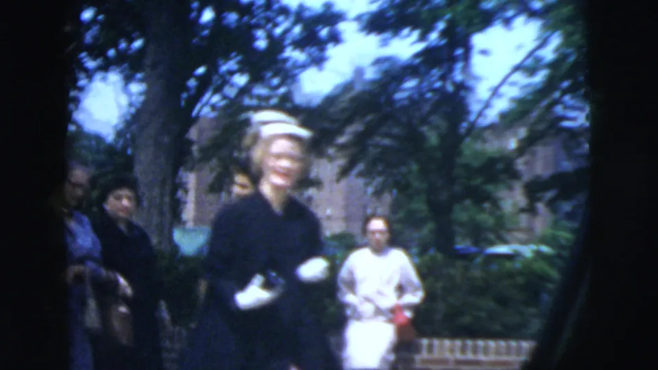 In 1958 New York City home-movie footage, a woman in a white hat and dark dress walks along a shaded residential sidewalk.