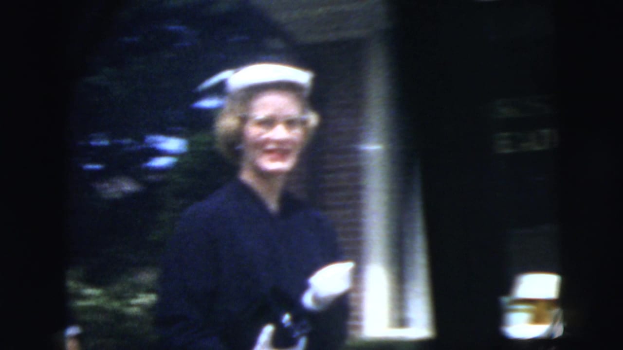 The woman in a white hat and dark dress smiles as she walks past a brick residential building in the supplied New York City setting.