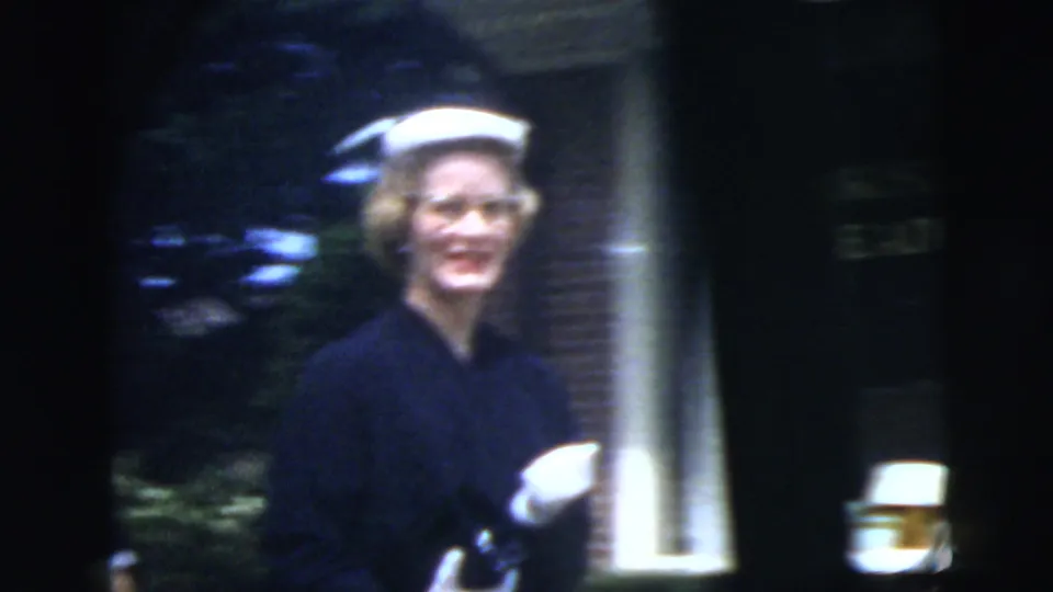 The woman in a white hat and dark dress smiles as she walks past a brick residential building in the supplied New York City setting.