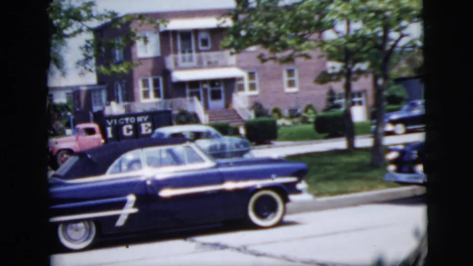 A residential New York City street view shows mid-century parked cars, brick apartment buildings, trees, and visible sign or vehicle text.