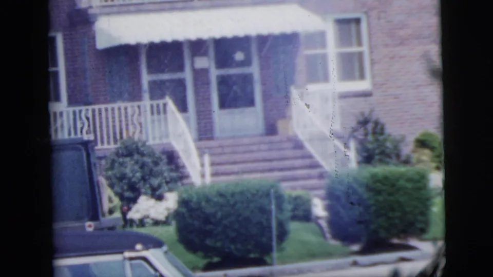1958 New York City home-movie frame showing a brick residential entrance, white awning, front steps, hedges and a parked car edge.
