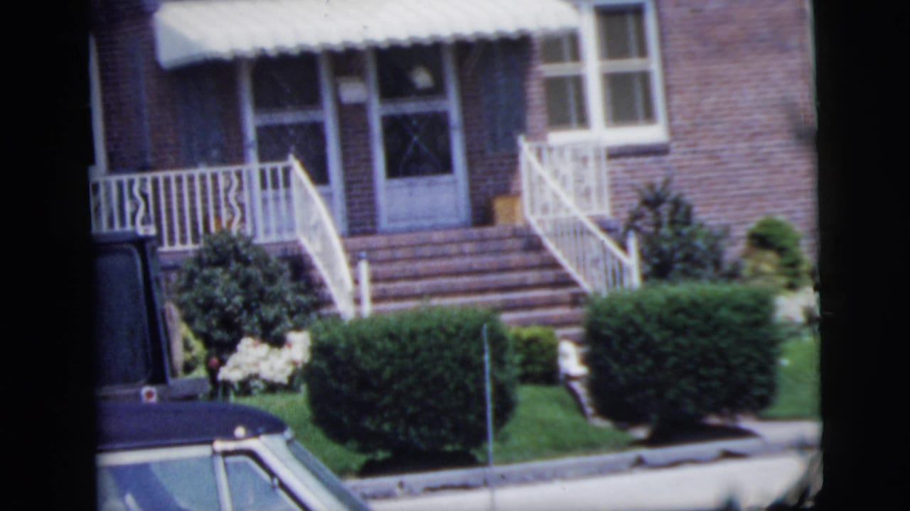 Brick house facade with white awning, steps, railings, hedges and curbside car roof.
