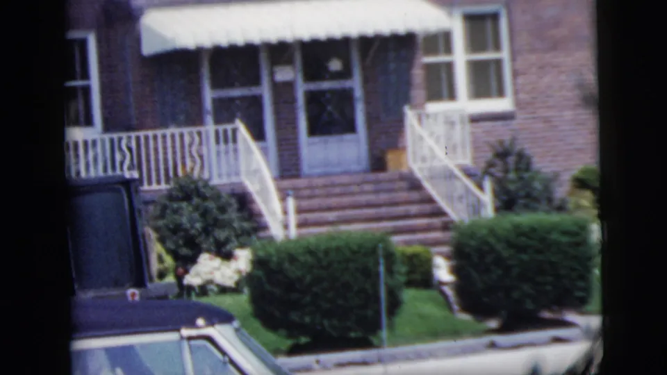 Closer 1958 New York City home-movie view of a brick house entrance with white awning, front steps, railings and curbside car.