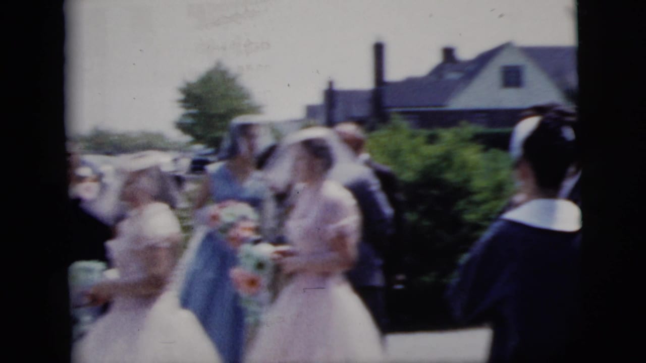 In 1958 New York City home-movie footage, a veiled bride and bridesmaids in pale dresses move past guests along a residential street.